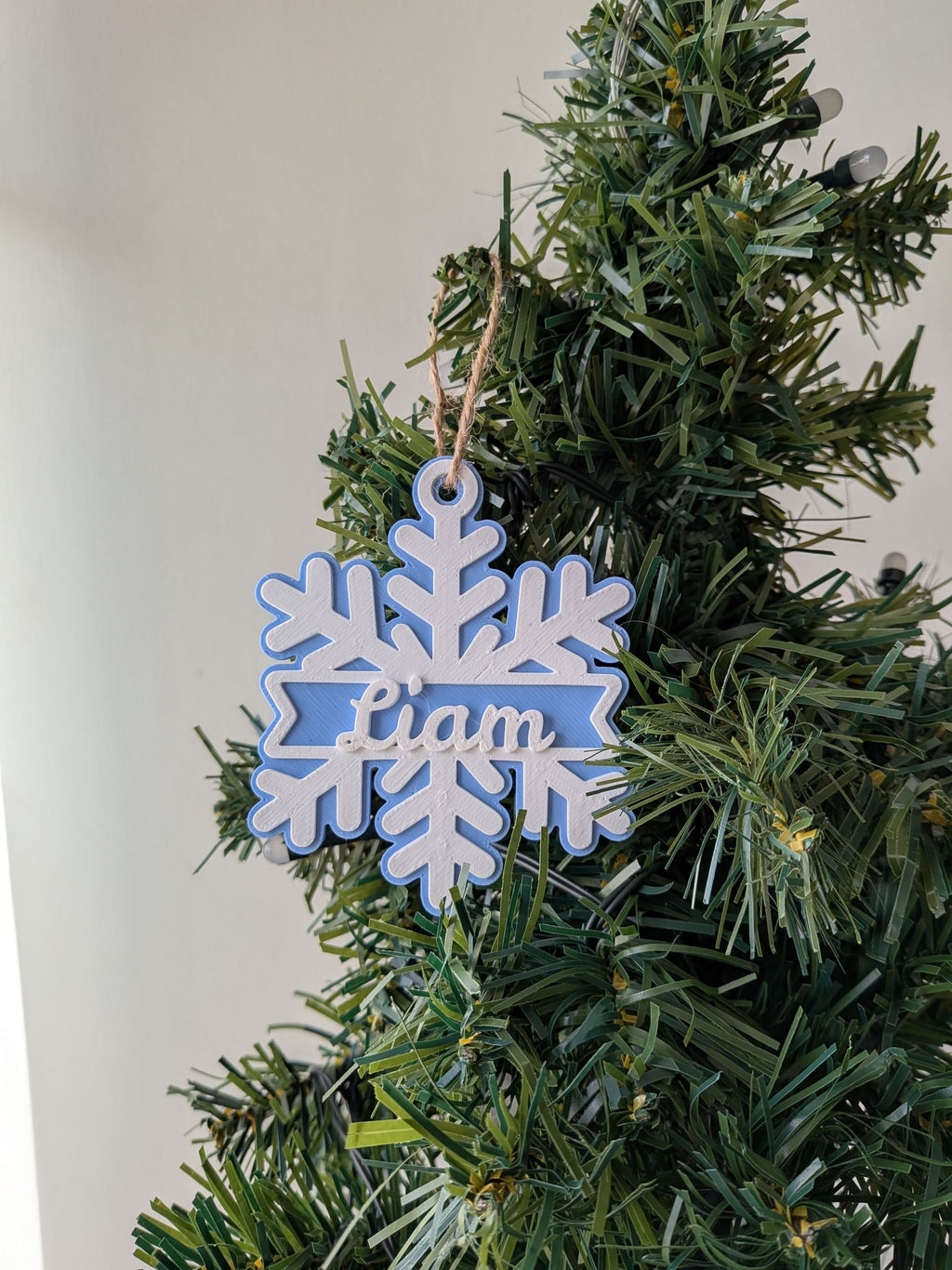 Flocon de Noël bleu clair et blanc personnalisé avec le prénom Liam, suspendu dans un sapin.